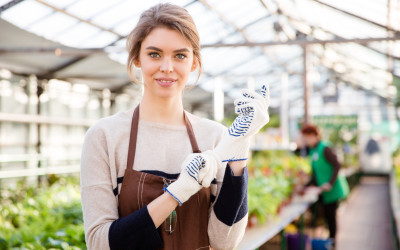 Happy young woman gardener in garden gloves and brown apron standing in greenhouse